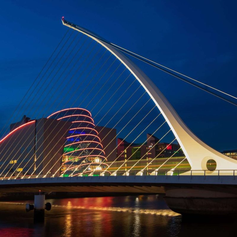 a bridge and some buildings along a river at night with lights in the water Samuel Beckett bridge and buildings illuminated in the light from the river's surface