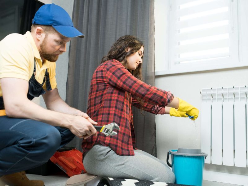 Serious attractive young lady in casual shirt and rubber gloves squeezing cloth while mopping floor from radiator water, repairman in cap sitting near her and waiting when he will start working